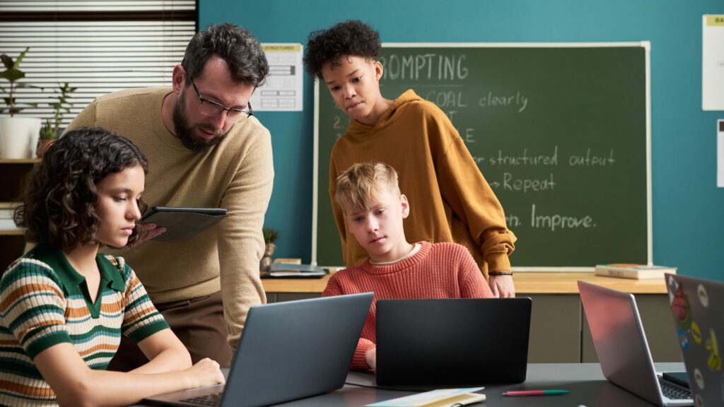 A teacher and three students work together at a table with laptops and a tablet, creating an engaging classroom learning environment surrounded by a chalkboard and educational materials.