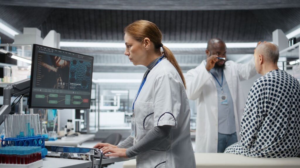 A female scientist works at a computer in a laboratory, contributing to clinical research, while two doctors examine a patient in the background. Test tubes and medical equipment are visible.