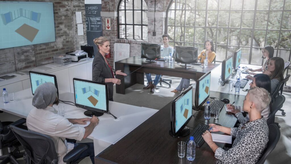 A woman from a local animation company stands and presents to a group of people seated at desks with computers in a modern office classroom; digital renderings are displayed on the monitors and screen.