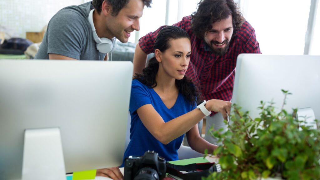 Three people collaborate at computer desks, with a woman pointing at a screen while two men look on; office plants and camera equipment highlight their creative workspace focused on Expedited Animation Services.