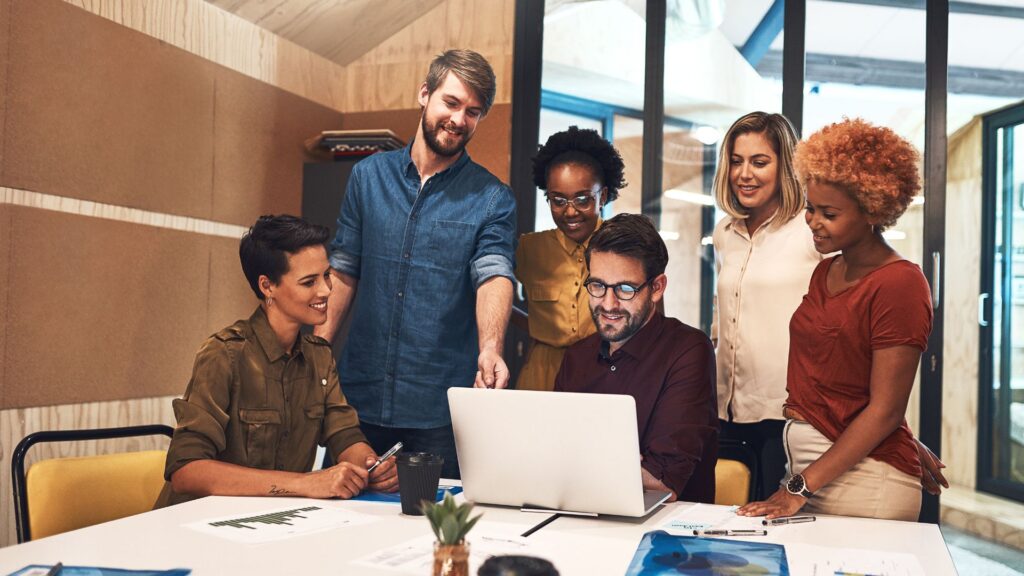 Six people gather around a table in a modern office, appearing engaged in discussion and collaboration as they review a Quick Turnaround Animation on a laptop screen.