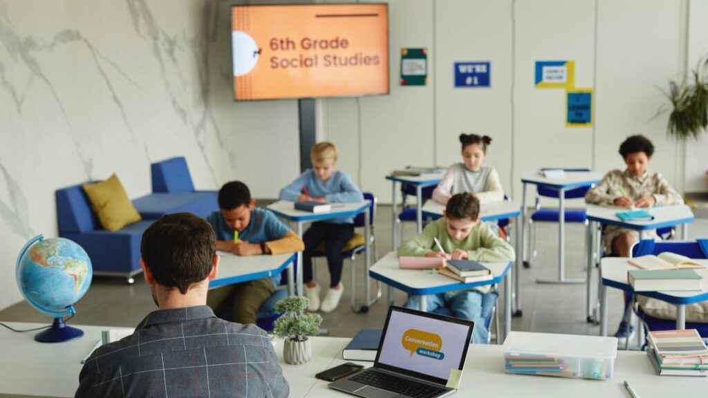A teacher sits at a desk facing students jotting notes in their notebooks during a 6th grade social studies class; a presentation is displayed on a screen, ideal for School Promotional Videos.