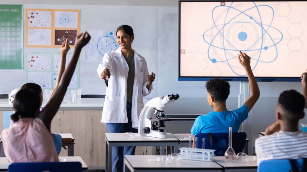 A teacher in a lab coat engages students in a science classroom, with a microscope on the desk and an atom diagram displayed on screen—perfect for School Promotional Videos showcasing active student participation.
