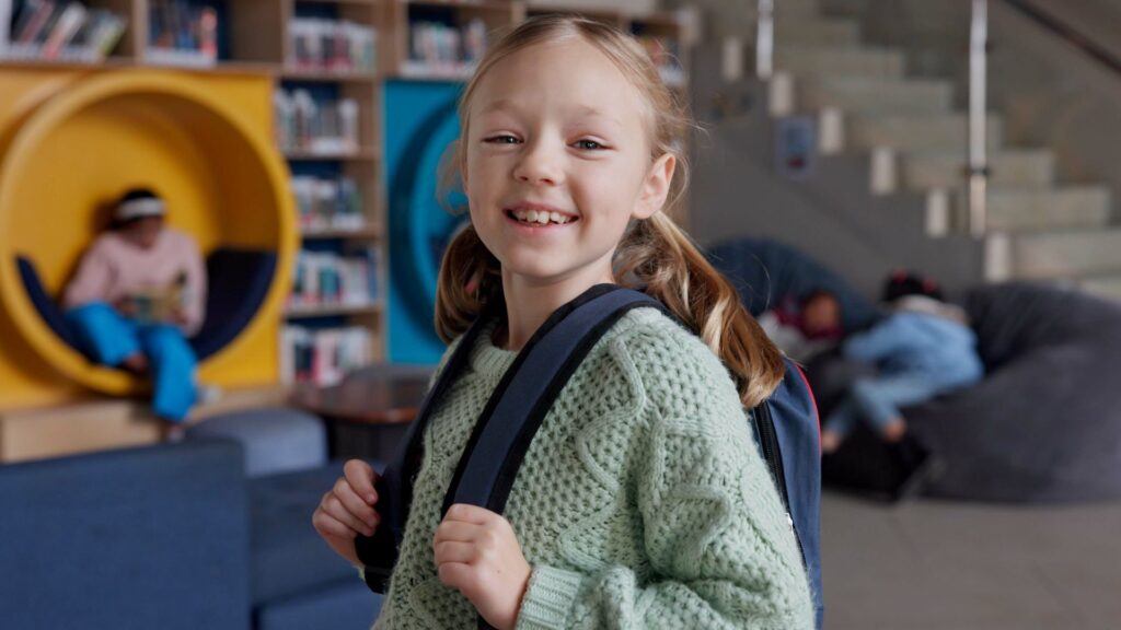 Smiling young girl with backpack stands in a library; bookshelves, children reading, and staircase are visible in the background—perfect for School Promotional Videos.