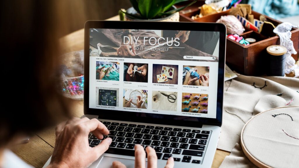 Person browsing a DIY website called "DIY Focus" on a laptop, with various craft projects and an engaging animation for website elements displayed on the screen, plus sewing supplies visible on the table.