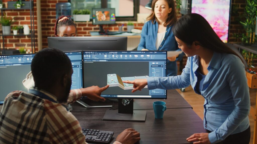 Four people collaborate at a desk with computers displaying 3D design software; two are pointing at a monitor and discussing the model, showcasing teamwork often found in top animation agencies.