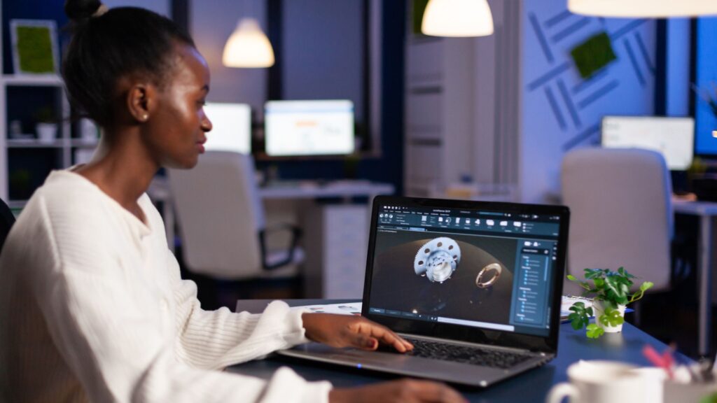 A woman sits at a desk using a laptop with 3D modeling software open, creating a digital model of a watch in an office—perfect for producing animation for YouTube channel content.