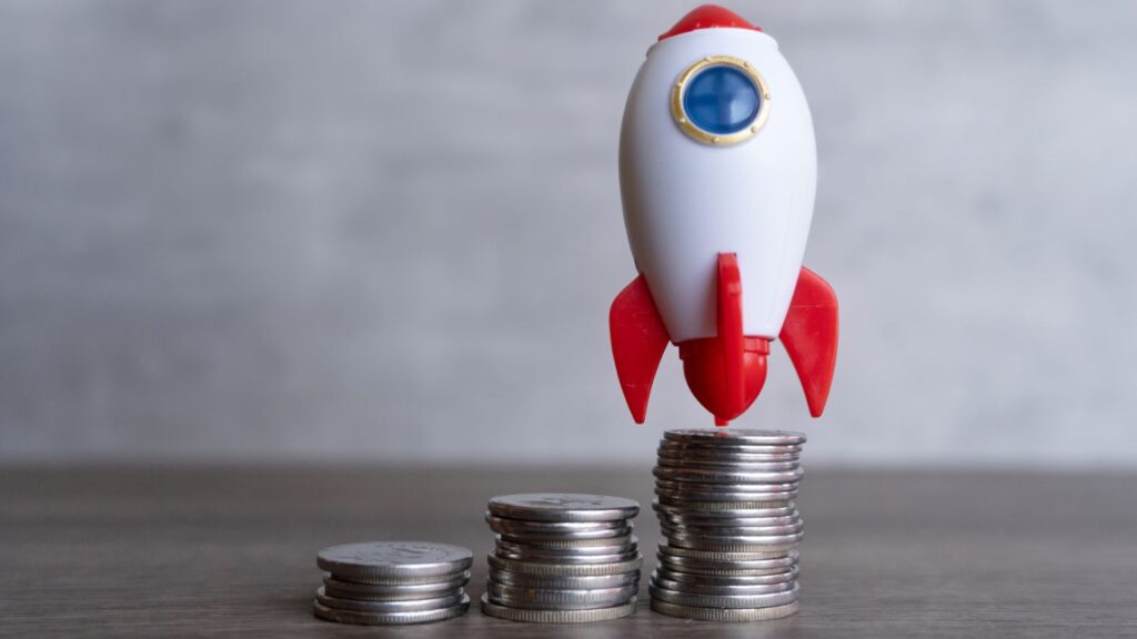 A small toy rocket, symbolizing educational animation funding, stands on top of stacked coins next to two shorter stacks against a plain background.