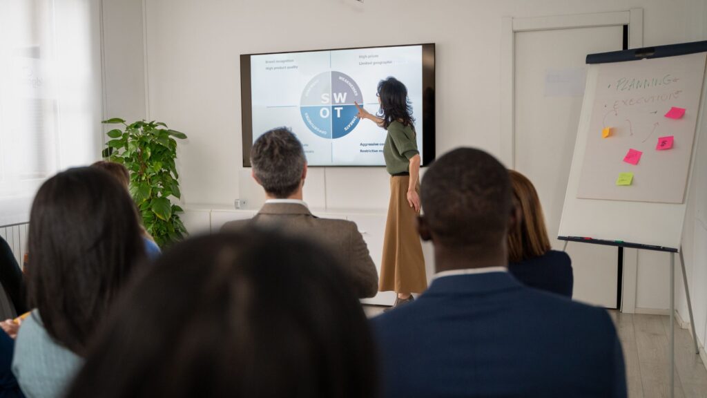 A woman presents a SWOT analysis on a screen to an audience in a meeting room, supported by Animated Presentation Services, with a flipchart and sticky notes visible in the background.
