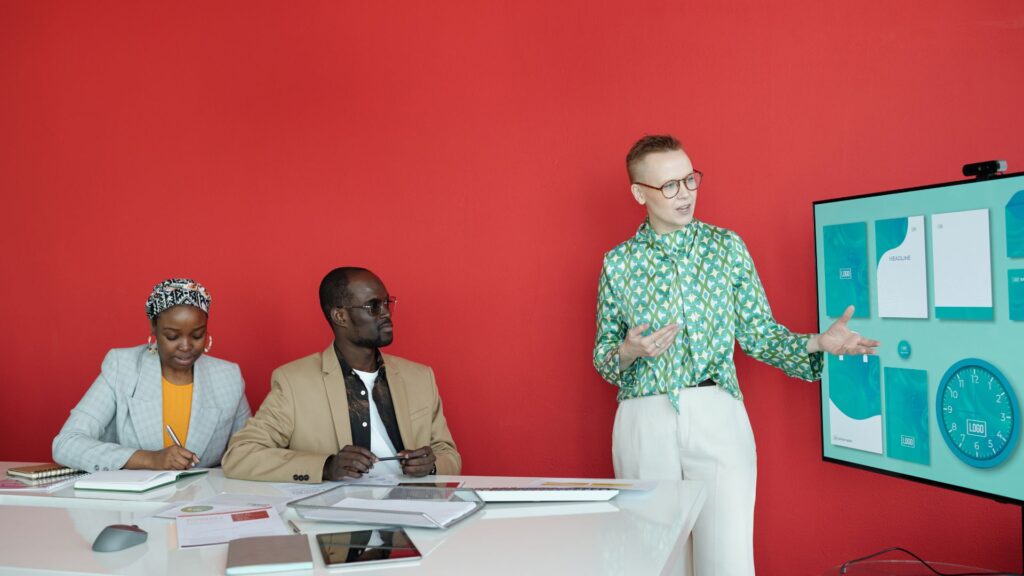 A person stands and gestures at an animated presentation on a screen while two colleagues sit at a desk taking notes, all in front of a bright red wall, showcasing the impact of Animated Presentation Services.