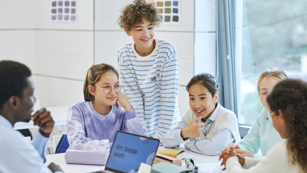 A group of students sits around a table in a classroom, talking and smiling, with a laptop labeled "6th Grade Math" and books in front of them, as an academic research animation plays on the screen.