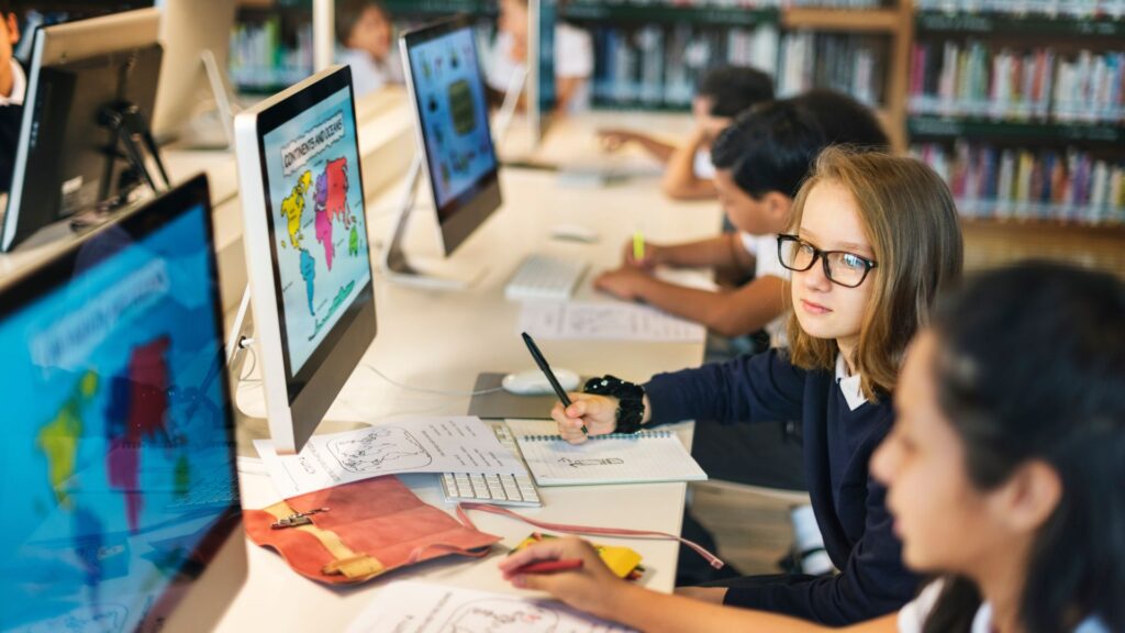 Students sit at a row of computers in a library, working on assignments involving maps and academic research animation. One student with glasses looks up while others remain focused on their work.