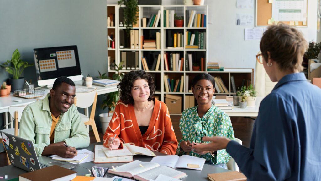 A group of three people sit at a table with books and notebooks, listening to a person standing and speaking in a modern office or classroom, exploring Educational Voice Alternatives.