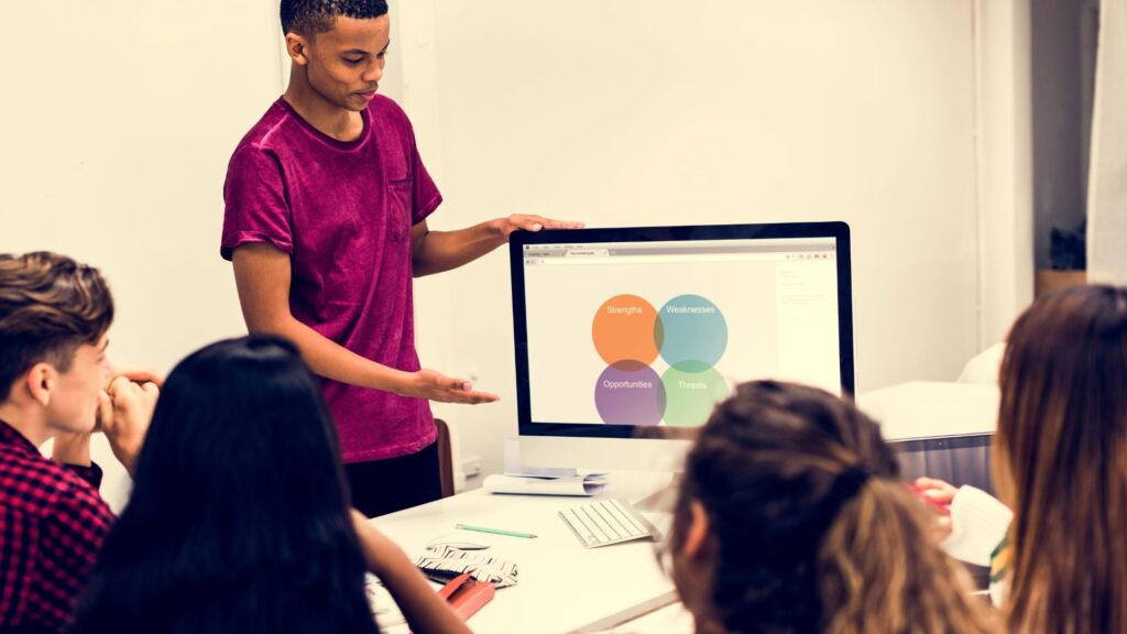 A person in a red shirt presents a Venn diagram displayed on a computer monitor to a group of seated colleagues, showcasing the benefits of Animated Presentation Services in a meeting room.