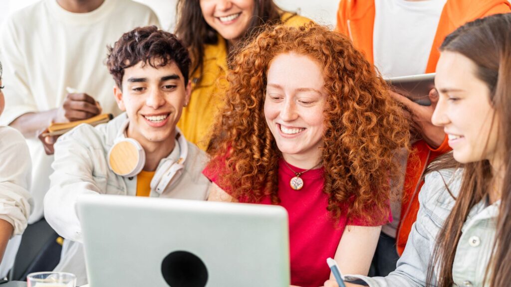 A group of young adults sit together around a laptop, smiling and collaborating on a project while exploring Educational Voice Alternatives.