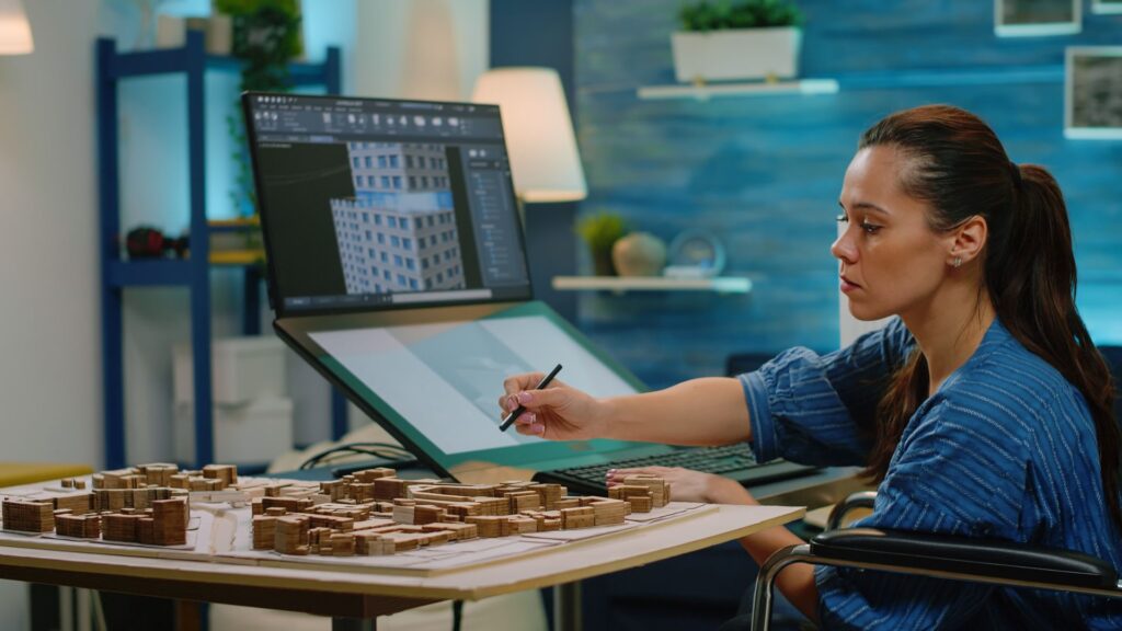 Woman sits at a desk working on an architectural model with small wooden structures, using a digital stylus and large monitor displaying a building design, enhanced by SME Animation Services for realistic visualizations.