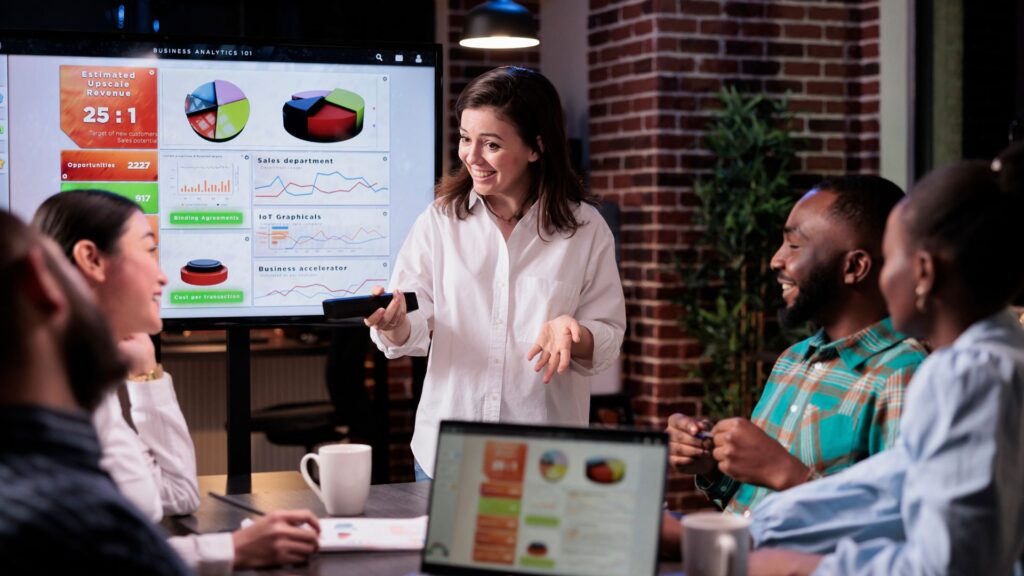 A woman presents business analytics data on a screen to four colleagues in a modern office meeting room, highlighting the impact of Animated Presentation Services.