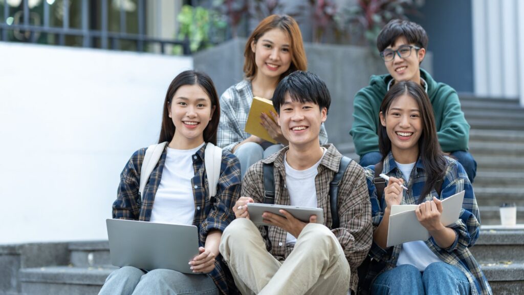 Five young adults sit on outdoor steps, smiling and holding electronic devices, notebooks, and pencils, engaging in academic research or animation projects together.