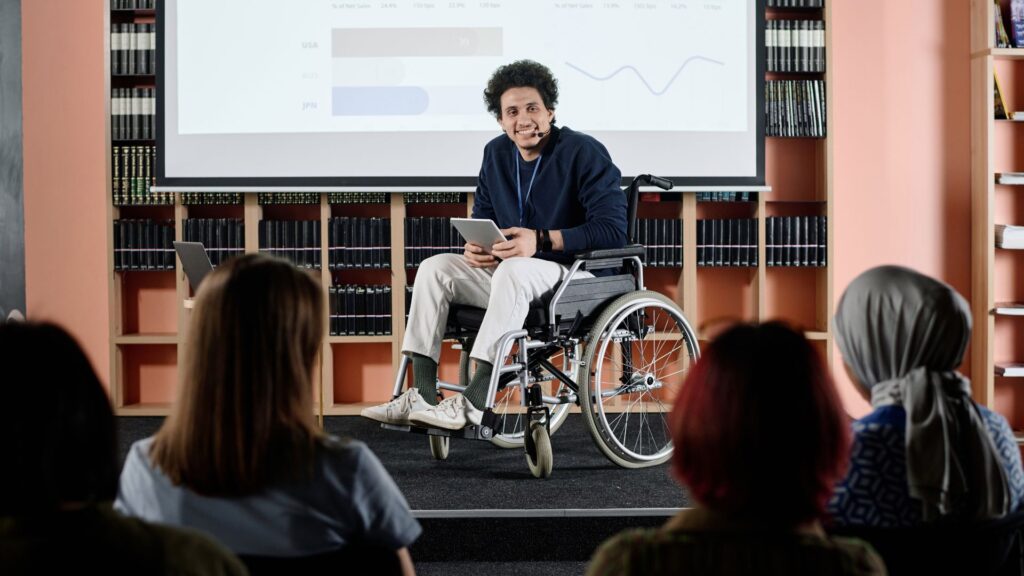A man in a wheelchair delivers a presentation with charts displayed on a screen, showcasing Animated Presentation Services to an audience seated in front of him in a library setting.