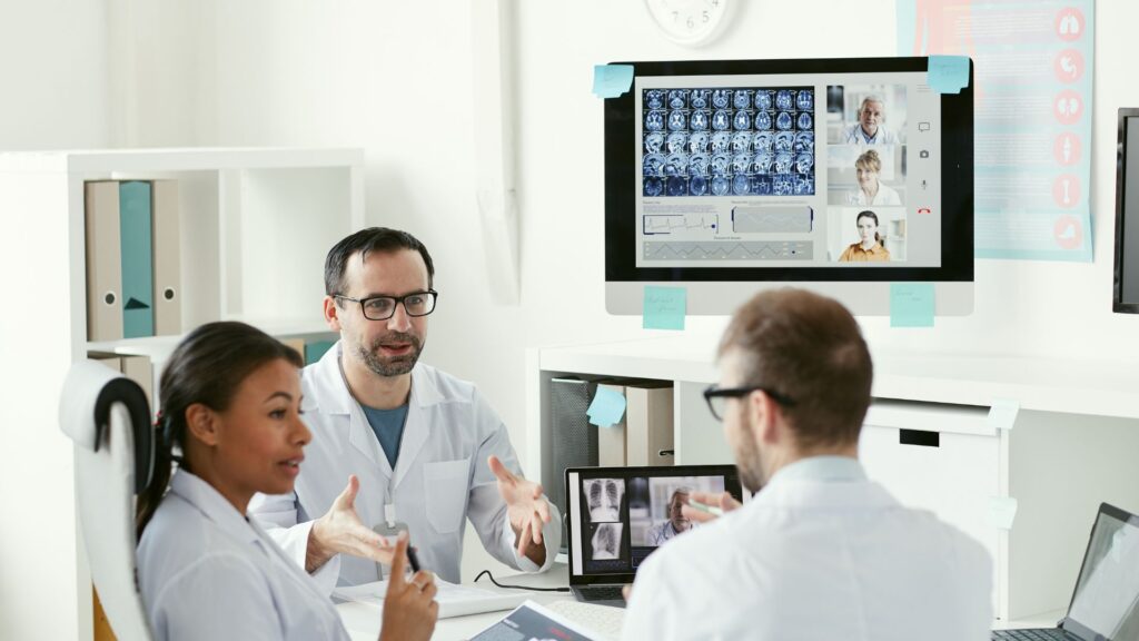 Three doctors in lab coats discuss patient scans and brain images displayed on a large monitor and a laptop in a modern medical office, highlighting the impact of Healthcare Marketing Animation in presenting complex data.