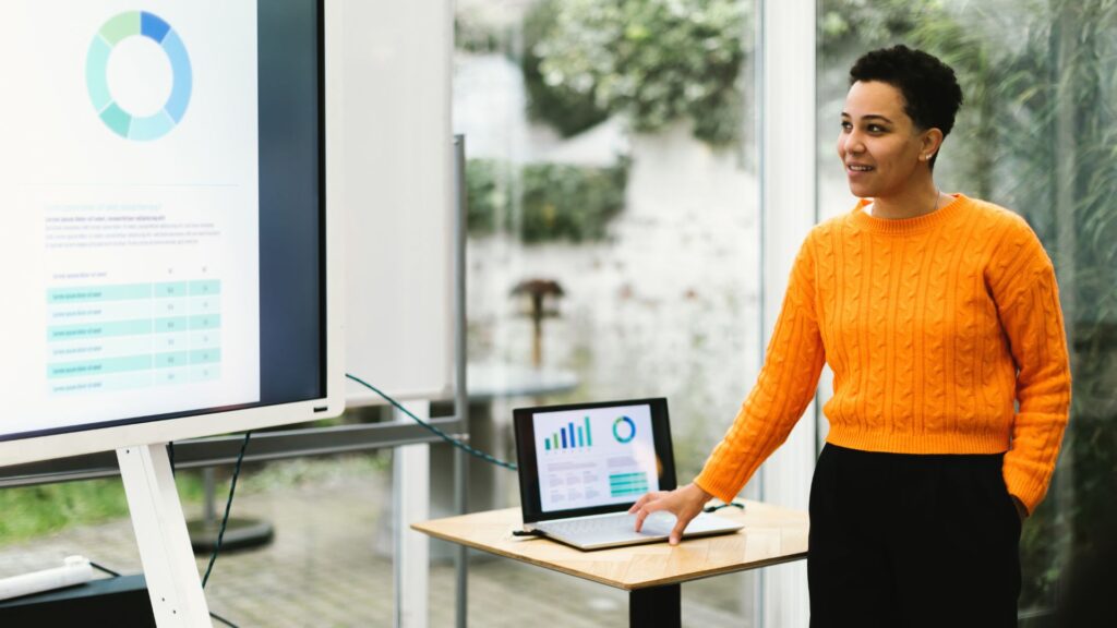 A person in an orange sweater uses Animated Presentation Services to showcase data visualizations on a large screen and a laptop in a bright room.