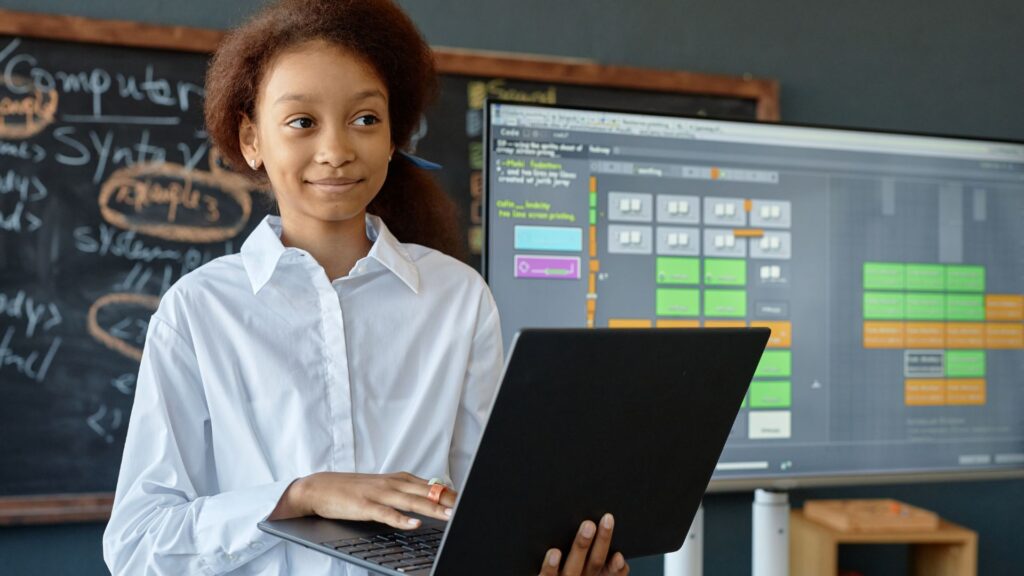 A student holding a laptop stands in a classroom with a chalkboard and a digital programming interface displayed on a screen in the background, exploring Educational Voice Alternatives.