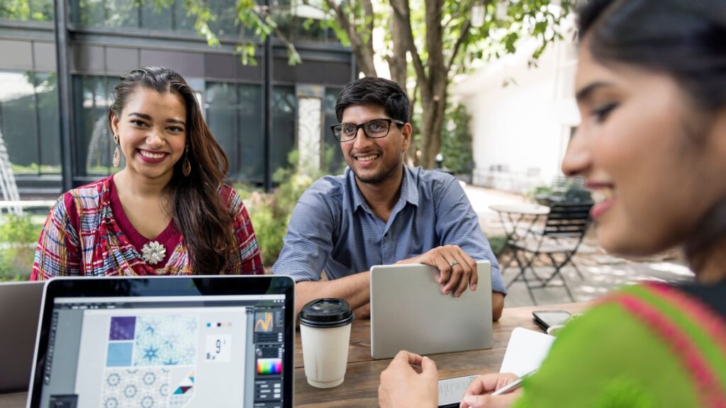 Three people sit at a table outdoors with laptops and notebooks, smiling and talking about Animation For YouTube Channel ideas, with trees and a building in the background.