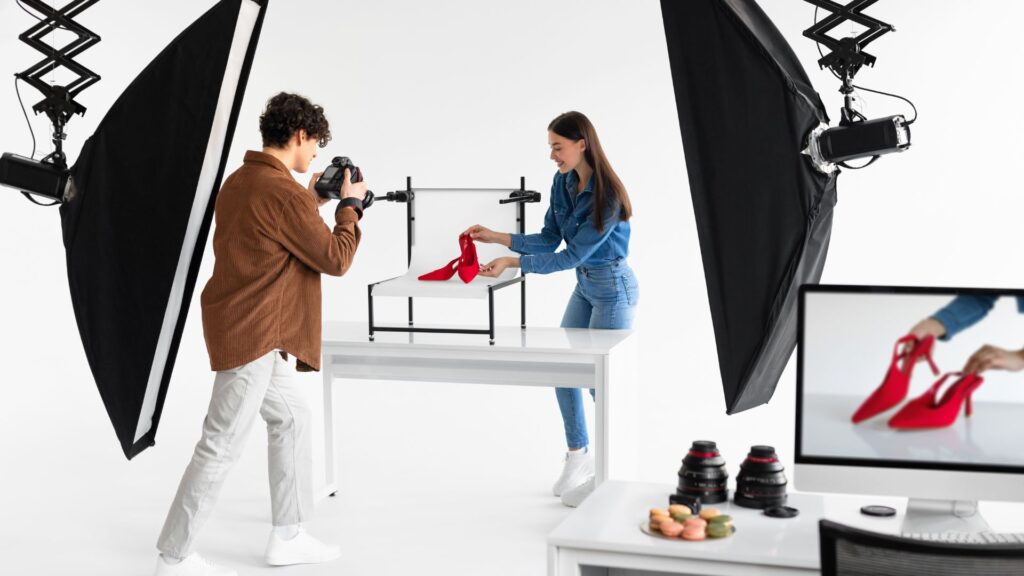 A photographer takes pictures of a woman holding a red shoe in a studio setup with professional lighting and camera, capturing shots for a product explanation animation. A computer monitor and camera lenses are visible on a desk nearby.