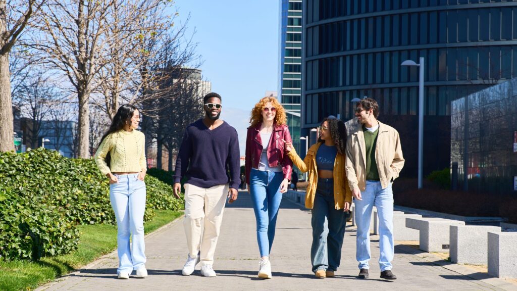 Five people are walking together on a sunny day along a paved path in an urban area, smiling and talking, as if enjoying a Virtual Campus Tour.