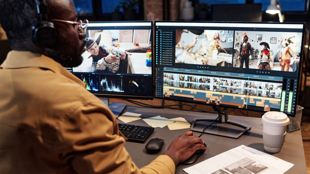 A person wearing headphones edits a Government Animation Tender video on dual monitors at a desk with a keyboard, mouse, papers, and a takeaway coffee cup.