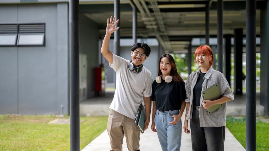 Three young adults with laptops and headphones stand outside a building; one waves at the camera while the other two smile, ready to explore their university with help from a virtual guide.