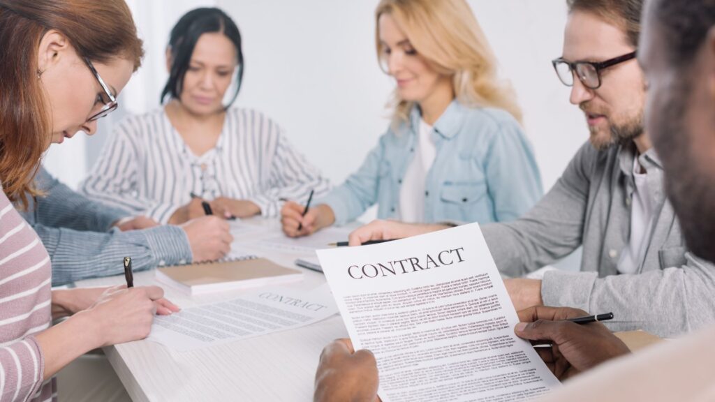 Five people sit at a table, reviewing and signing documents labeled "CONTRACT" for a Government Animation Tender, with pens and papers in front of them.