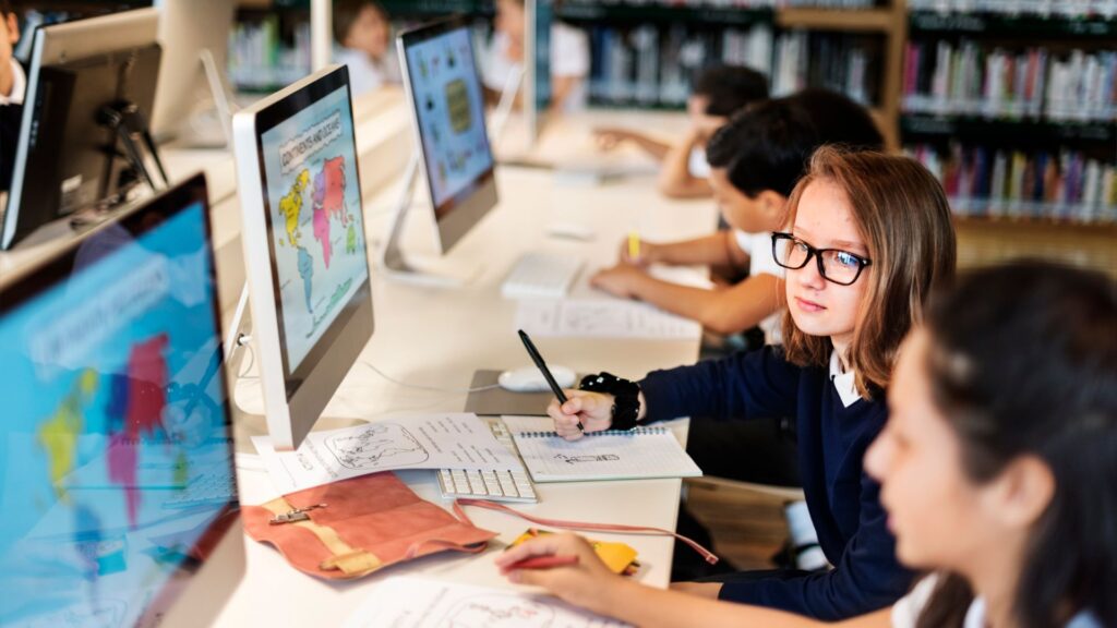 Students sit at a desk with desktop computers in a library, working on assignments with papers and notebooks in front of them. One student looks toward the camera, possibly exploring animation for an eLearning platform as part of their studies.