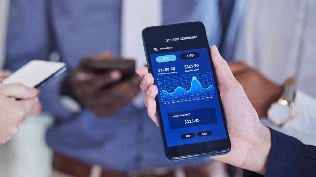A person holds a smartphone displaying a cryptocurrency trading app with BTC and USD prices, featuring modern visuals, while other people use their phones in the background.