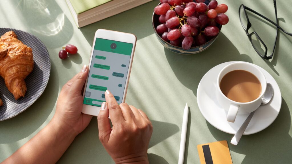 A person uses a smartphone with a chat and financial app at a table set with a croissant, grapes, coffee, a pen, and credit card, surrounded by modern visuals and subtle app animation on the screen.