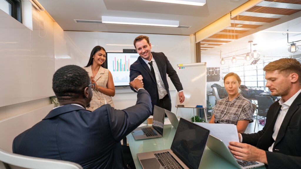 Four people in business attire meet in a conference room with laptops; two men fist bump, a woman claps, and a screen displays a bar chart showcasing their success on a recent Government Animation Tender.