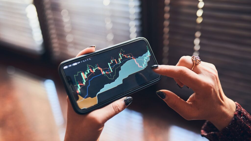 Person holding a smartphone displaying a candlestick stock chart with technical indicators in a sleek financial app, touching the screen to interact with its modern visuals.