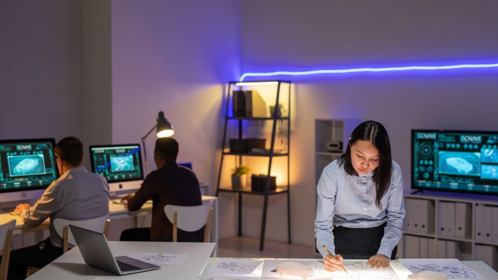 A woman reviews documents at a desk while two people work on computers displaying technical diagrams in a modern office with blue ambient lighting, reminiscent of animation studios like Educational Voice.