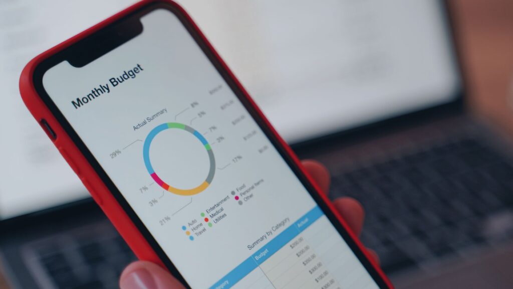A smartphone displays a monthly budget chart with modern visuals, featuring a pie graph and category breakdown, held in front of a blurred laptop screen.