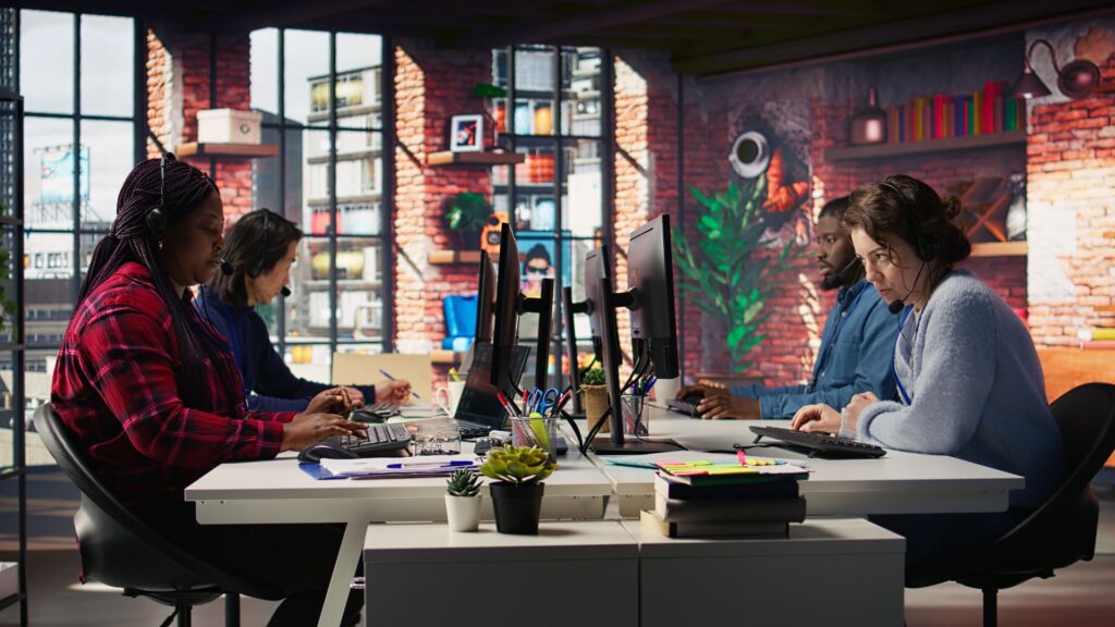 Four people work at computers in a modern office with large windows and brick walls, resembling animation studios like Educational Voice, with various office supplies on their desks.