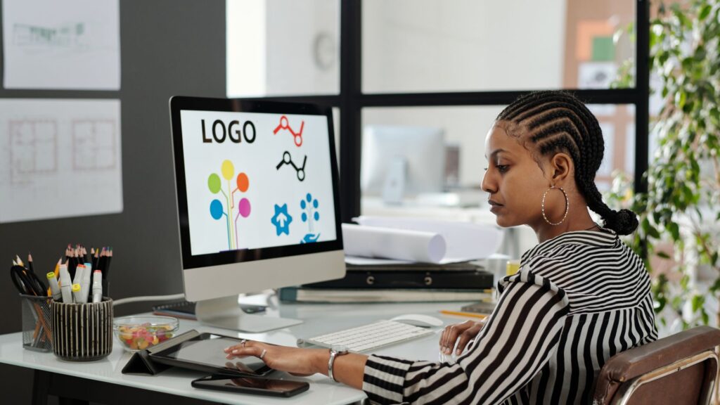 A woman sits at a desk in Ireland, working on a desktop computer displaying various brand identity and animated logo design concepts.
