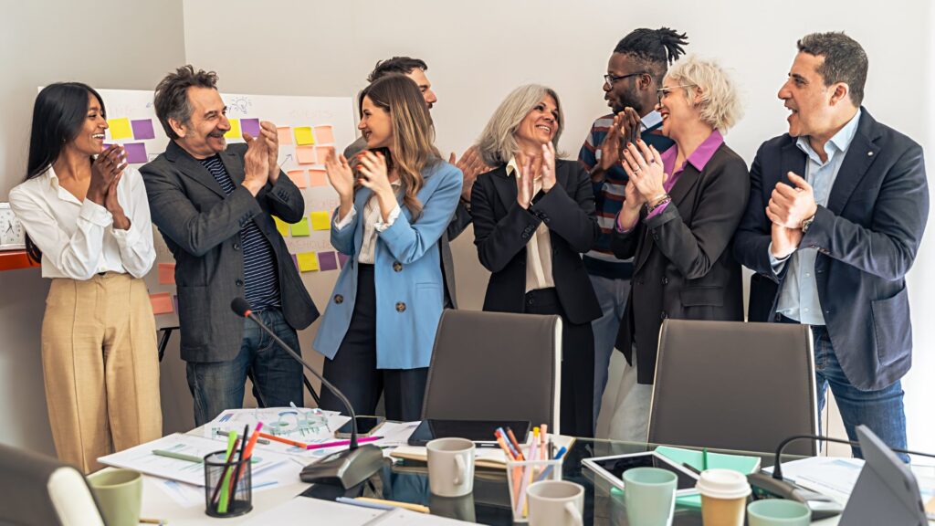 A diverse group of people stand in an office, smiling and applauding each other beside a whiteboard covered with colorful sticky notes highlighting ideas for upcoming Nonprofit Animation Grants.