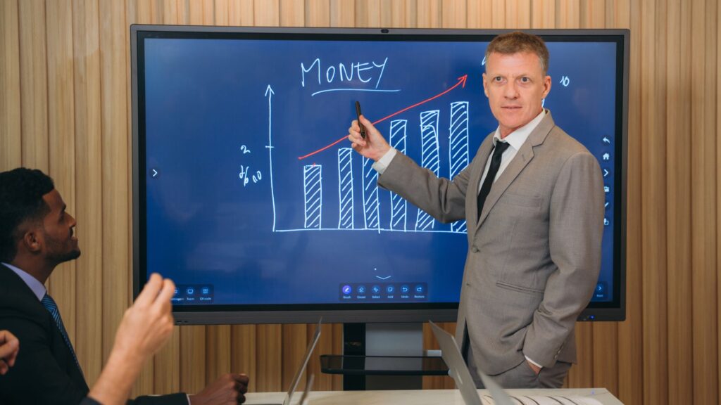 A man in a suit points to a bar graph labeled "MONEY" on a large screen during a business presentation, using animation to enhance financial education for his colleagues seated nearby.