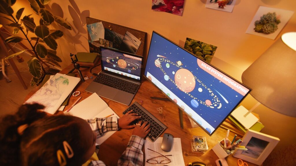 A person sits at a desk looking at a solar system diagram on a computer monitor and laptop, surrounded by notebooks, stationery, and plants—an inspiring scene reminiscent of animation studios like Educational Voice.