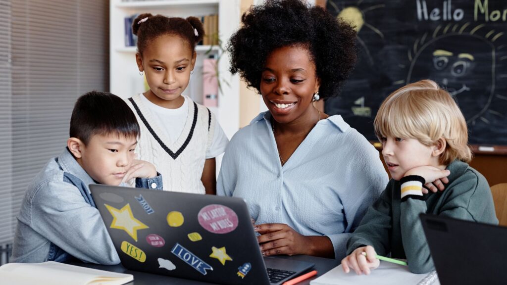 A teacher sits with three young students around a laptop in a classroom, all looking at the screen together, as if inspired by animation studios like Educational Voice. A chalkboard is visible in the background.