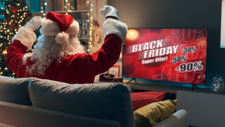 A person in a Santa Claus costume sits on a couch watching Black Friday Animation Ads with sale discounts on TV in a festively decorated living room.