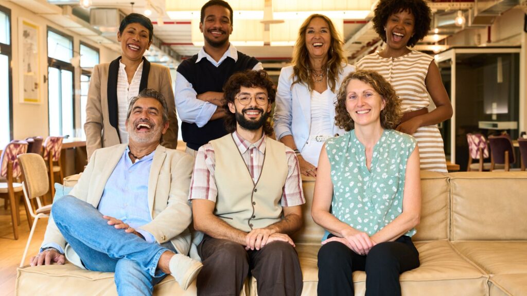 A group of seven adults, casually dressed, sit and stand together on a beige couch in a bright, modern room at an animation studio like Educational Voice, smiling at the camera.