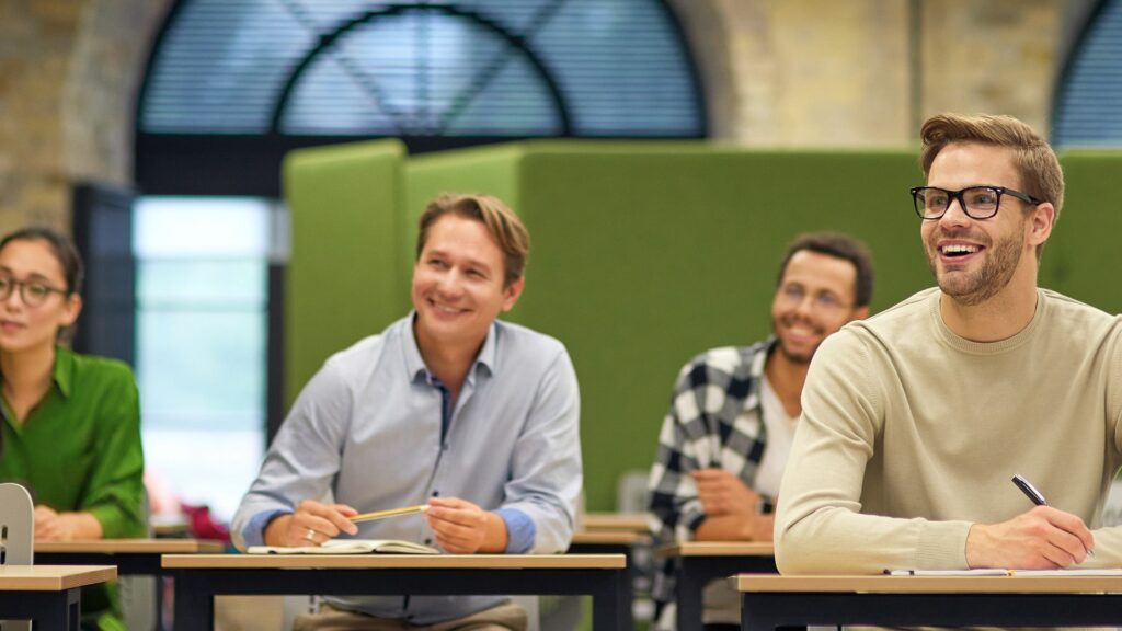 Four adults sit at desks in a classroom, attentively facing forward and smiling, with notebooks and pencils in front of them, as they engage in a lively discussion about effective university marketing strategies.