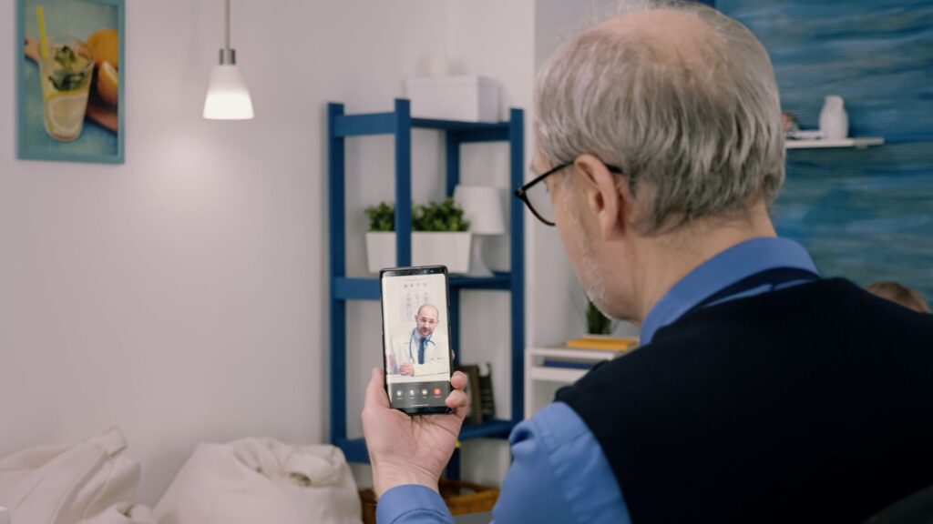 An older man with gray hair and glasses has a video call with a doctor on his smartphone in a well-lit, modern living room, illustrating the high impact of modern healthcare explainer videos.