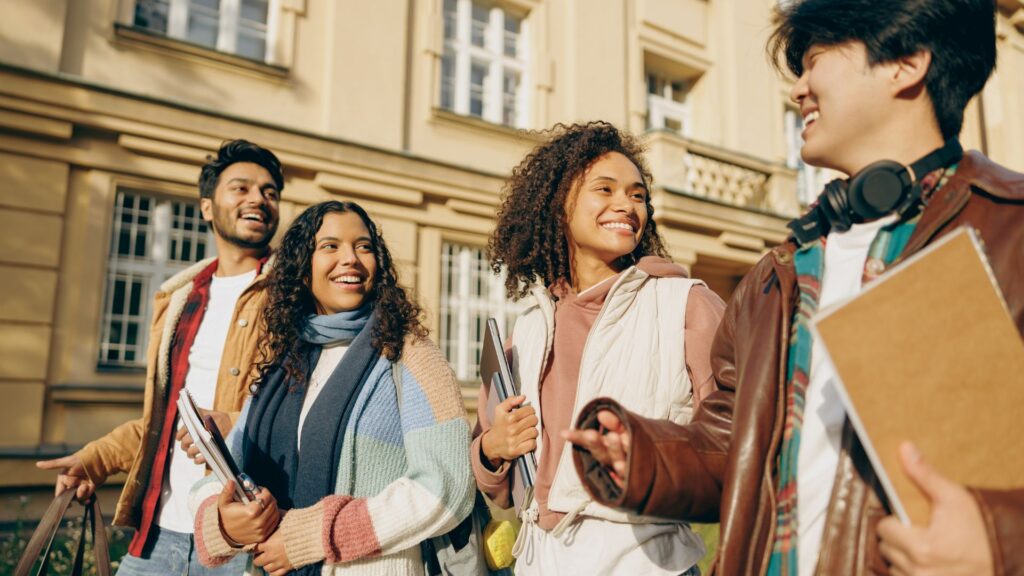 Four students walk together outside a school building, holding books and smiling as they discuss marketing strategies for their university project.
